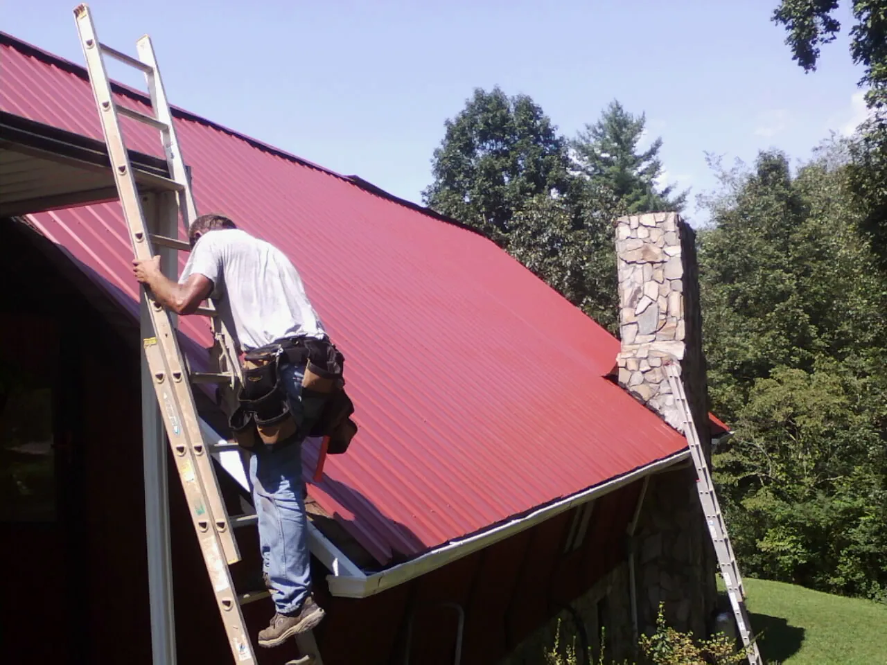 Roofer on a ladder beside a red metal roof and stone chimney — Soffit & Fascia Repair by Roofing Brothers in Vernon Center